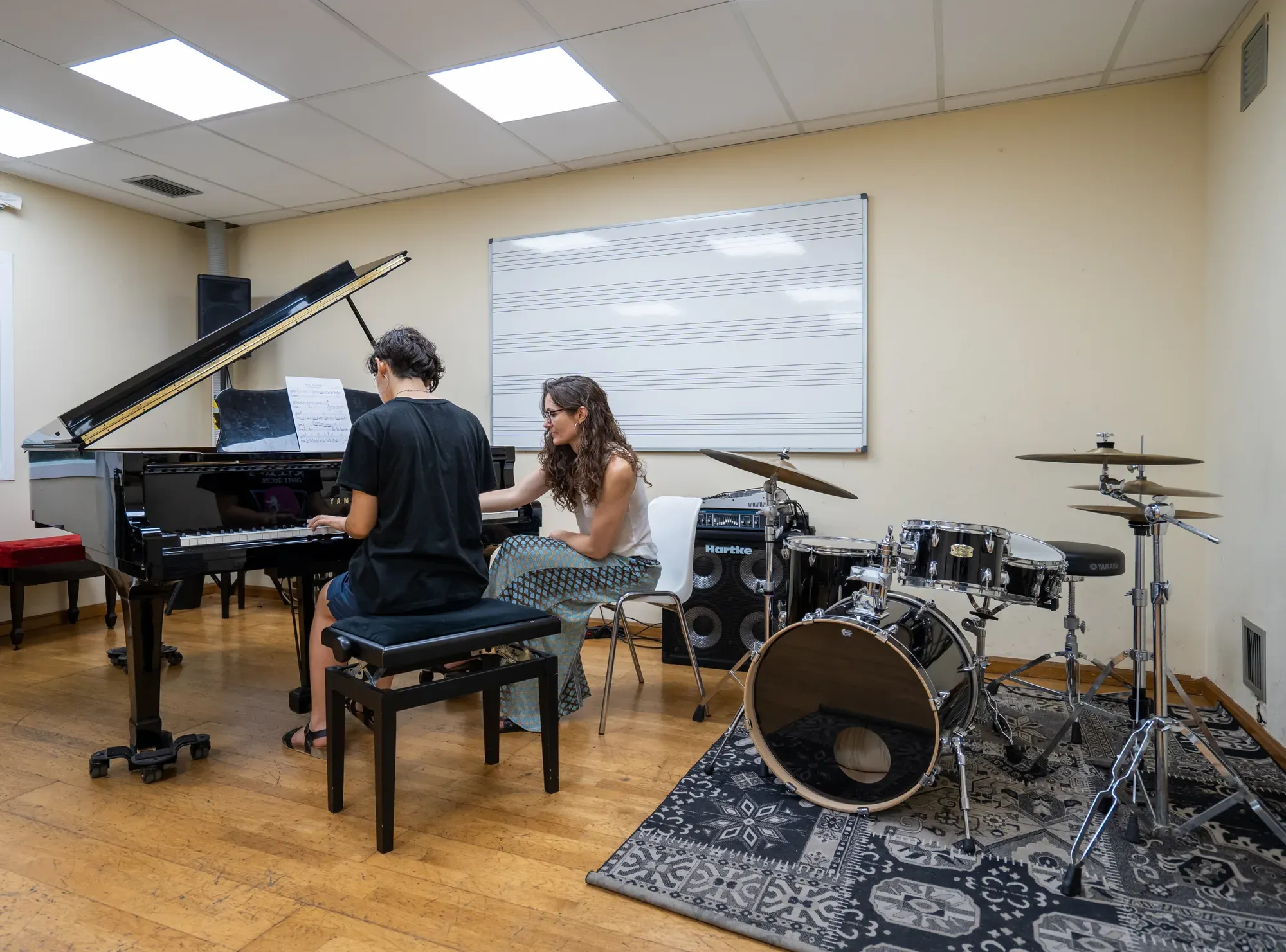 Un alumne al piano amb la professora en una aula amb bateria